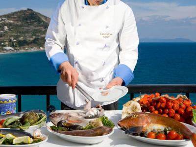 Chef prepara pesce fresco con vista mare, colori vivaci e atmosfera mediterranea.