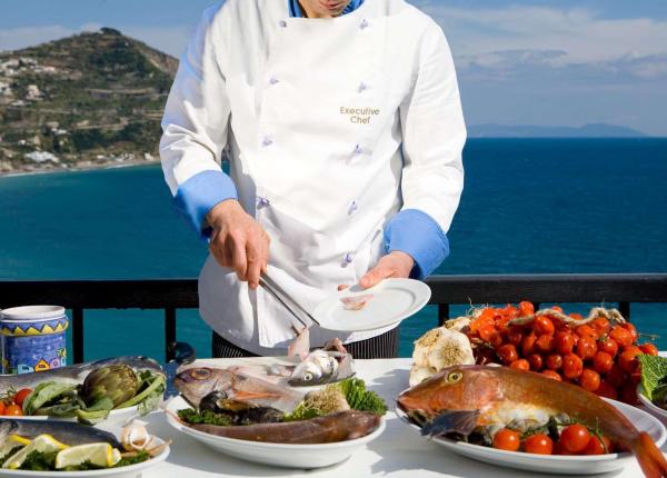 Chef prepara pesce fresco con vista mare, colori vivaci e atmosfera mediterranea.